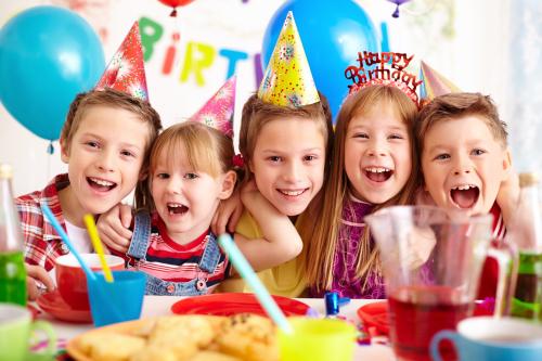 Deca sa rodjendanskim kapama na glavi ispred stola punog hrane i pića, smeju se i raduju